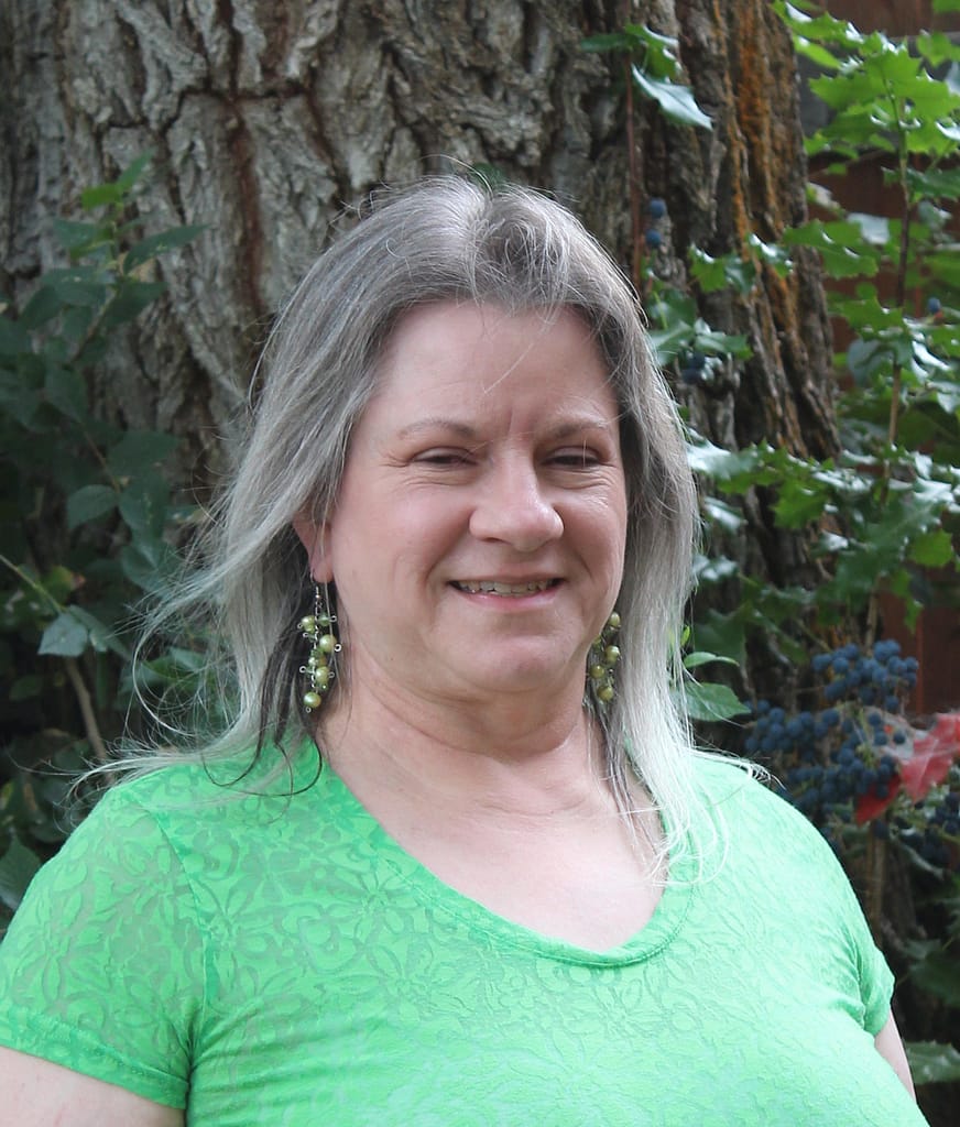 Photo of Pam VanDerDoes standing in front of a tree and smiling at the camera. Her wavy brown and silver hair is down over her shoulders. She is wearing a green top and green earrings.